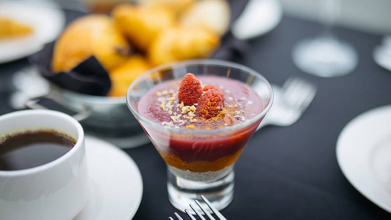 Un dessert élégant dans un verre, composé de couches de mousse ou de purée de fruits rouges, garni de framboises fraîches et de miettes de noix ou de biscuits, accompagné d'une tasse de café et de croissants en arrière-plan.