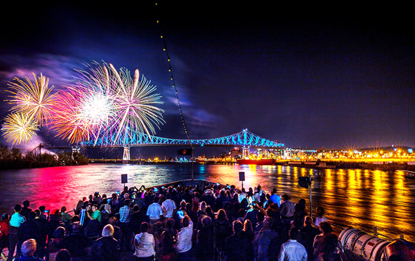 Une foule profite d’un feu d’artifice vibrant depuis la terrasse principale du AML Cavalier Maxim à Montréal avec vue sur le Pont Jacques Cartier et la ville illuminés en arrière plan.