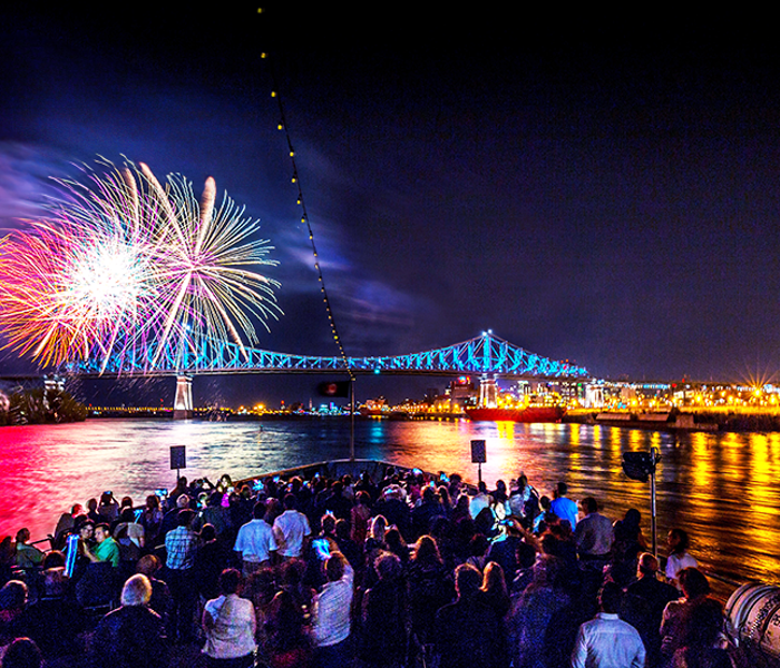 Une foule profite d’un feu d’artifice vibrant depuis la terrasse principale du AML Cavalier Maxim à Montréal avec vue sur le Pont Jacques Cartier et la ville illuminés en arrière plan.