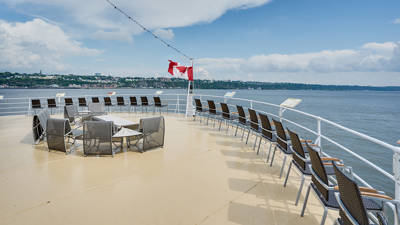 Terrasse extérieure avant du navire AML Louis Jolliet à Québec avec des rangées de chaises et de tables, un drapeau canadien flottant, avec vue sur le fleuve Saint-Laurent