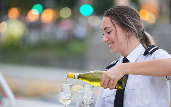 Une serveuse souriante en uniforme verse du vin blanc dans des verres, avec un arrière-plan flou de lumières et de verdure.