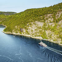 Aerial view of a ferry cruising through a winding, forested river. The vessel leaves a trail in the deep blue water, with rocky cliffs and dense green trees lining the riverbanks under a clear sky.