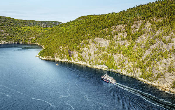 Vue aérienne d'un ferry naviguant à travers une rivière sinueuse bordée de forêts. Le navire laisse une traînée dans l'eau bleu profond, tandis que des falaises rocheuses et des arbres denses et verdoyants longent les rives sous un ciel clair.