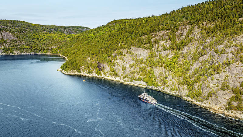 Vue aérienne d'un ferry naviguant à travers une rivière sinueuse bordée de forêts. Le navire laisse une traînée dans l'eau bleu profond, tandis que des falaises rocheuses et des arbres denses et verdoyants longent les rives sous un ciel clair.