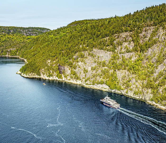 Vue aérienne d'un ferry naviguant à travers une rivière sinueuse bordée de forêts. Le navire laisse une traînée dans l'eau bleu profond, tandis que des falaises rocheuses et des arbres denses et verdoyants longent les rives sous un ciel clair.