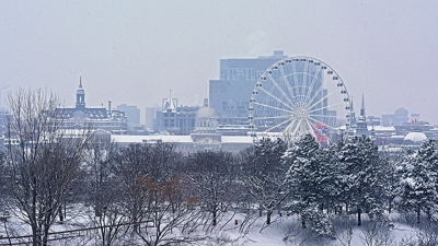 A view of snowy Montreal with the ferris wheel amidst snow-covered trees in the foreground and the city skyline in the background.