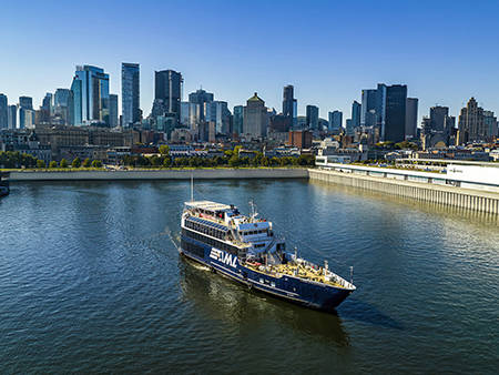 Un bateau de croisière naviguant sur une rivière, avec une vue panoramique sur une ville avec de hauts gratte-ciels en arrière-plan sous un ciel dégagé.