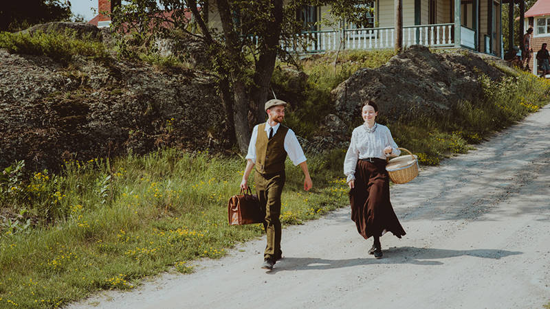 Deux personnages habillés en tenue d'époque marche sur Grosse Île. Ils descendent une petite pente. Il y a une maison d'époque en arrière plan 