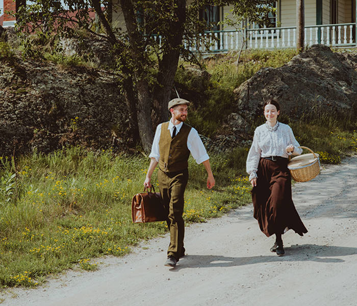 Deux personnages habillés en tenue d'époque marche sur Grosse Île. Ils descendent une petite pente. Il y a une maison d'époque en arrière plan 