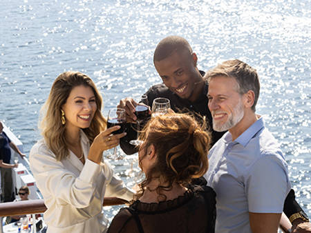  A group of smiling friends toasting with glasses of red wine on a boat deck, under bright sunlight, with a view of the water in the background.