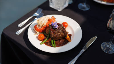 Une assiette gastronomique comprenant un filet de viande nappé de sauce, accompagné de légumes verts, tomates cerises, purée et garniture de fleurs comestibles, avec un homard grillé à côté. Le tout est présenté sur une nappe noire avec des couverts élégants.