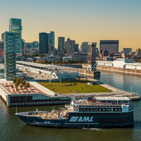 A blue cruise ship with the "AML" logo sails near a modern quay with green spaces, surrounded by the skyscrapers of a city under a clear and sunny sky.