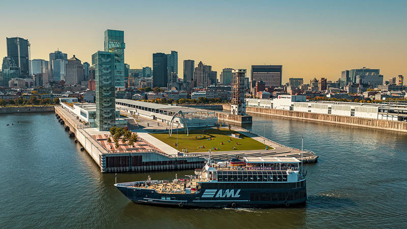 Un bateau de croisière bleu avec le logo "AML" navigue près d'un quai moderne avec des espaces verts, entouré par les gratte-ciels d'une ville sous un ciel clair et ensoleillé.