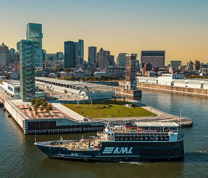 Un bateau de croisière bleu avec le logo "AML" navigue près d'un quai moderne avec des espaces verts, entouré par les gratte-ciels d'une ville sous un ciel clair et ensoleillé.