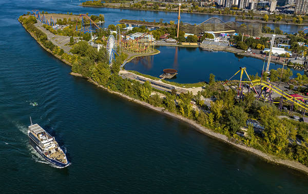 Vue aérienne d'une île avec un parc d'attractions comprenant des montagnes russes, une grande roue et des manèges, entourée d'une rivière. Un bateau de croisière navigue sur l'eau à gauche.