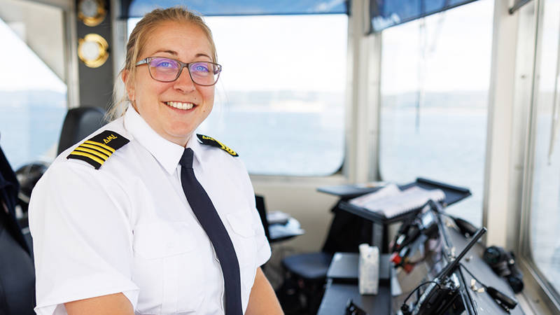 Une capitaine maritime en uniforme blanc, avec des insignes noirs et dorés sur les épaules, sourit sur la passerelle d’un navire. Elle porte des lunettes et a une sucette dans la bouche. Les commandes de la passerelle et les fenêtres offrant une vue sur l’océan sont visibles en arrière-plan.