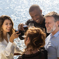 Un groupe d'amis souriants trinque avec des verres de vin rouge sur le pont d'un bateau, sous un soleil éclatant, avec une vue sur l'eau en arrière-plan.