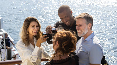 Un groupe d'amis souriants trinque avec des verres de vin rouge sur le pont d'un bateau, sous un soleil éclatant, avec une vue sur l'eau en arrière-plan.