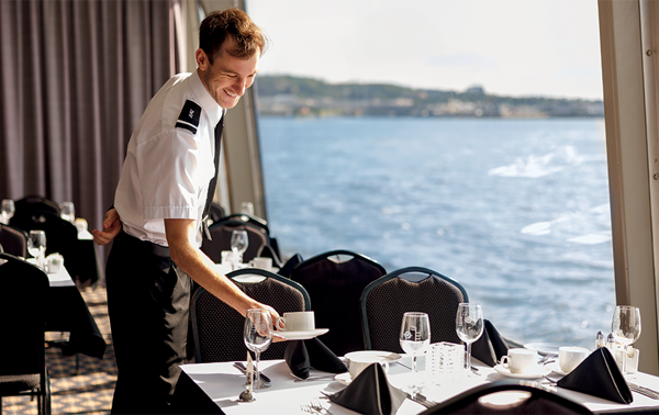 A smiling waiter in uniform setting a table near a large window overlooking the St. Lawrence River aboard the AML Cavalier Maxim in Montreal.