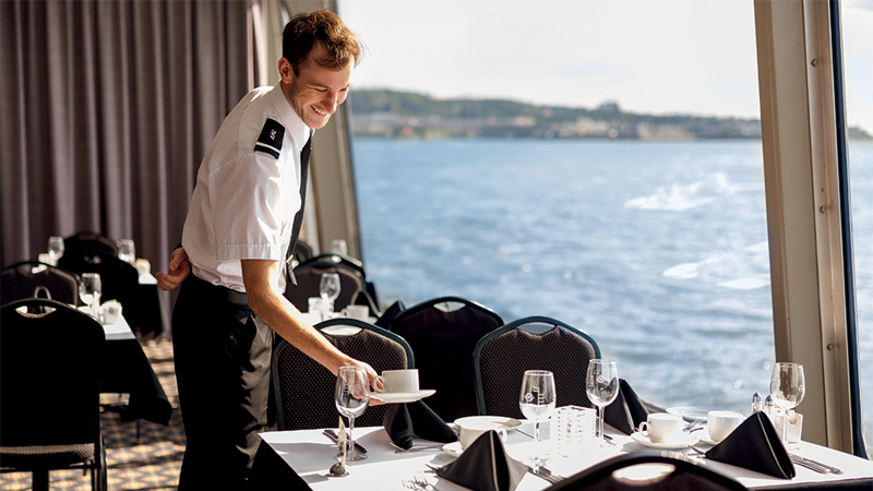 Un serveur souriant en uniforme dresse une table près d'une grande fenêtre donnant sur le fleuve Saint-Laurent à bord du navire AML Cavalier Maxim à Montréal.