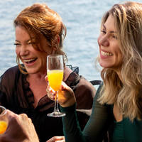 A group of friends enjoying a cheerful moment in a bright restaurant, with a smiling woman holding a glass of mimosa.