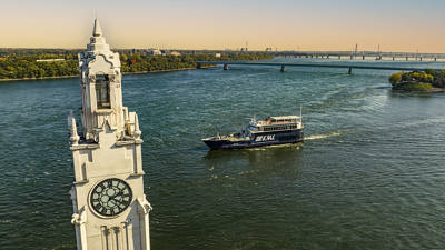 Vue aérienne d'une tour avec une grande horloge située au bord d'une rivière. Un bateau de croisière noir marquée "AML" navigue sur l'eau, avec un pont en arrière-plan et des rives verdoyantes.