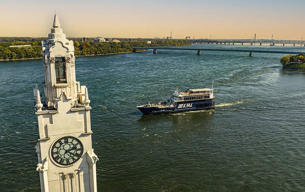 Vue aérienne d'une tour avec une grande horloge située au bord d'une rivière. Un bateau de croisière noir marquée "AML" navigue sur l'eau, avec un pont en arrière-plan et des rives verdoyantes.