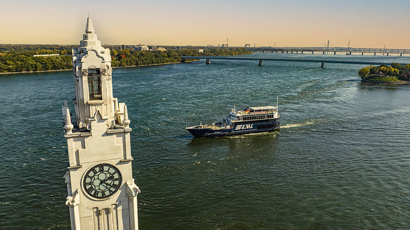 Vue aérienne d'une tour avec une grande horloge située au bord d'une rivière. Un bateau de croisière noir marquée "AML" navigue sur l'eau, avec un pont en arrière-plan et des rives verdoyantes.