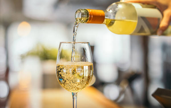 Un gros plan montre une main versant du vin blanc d'une bouteille dans un verre à vin. Le vin pétillant s'écoule doucement et remplit le verre, tandis que l'arrière-plan flou évoque une atmosphère chaleureuse et conviviale