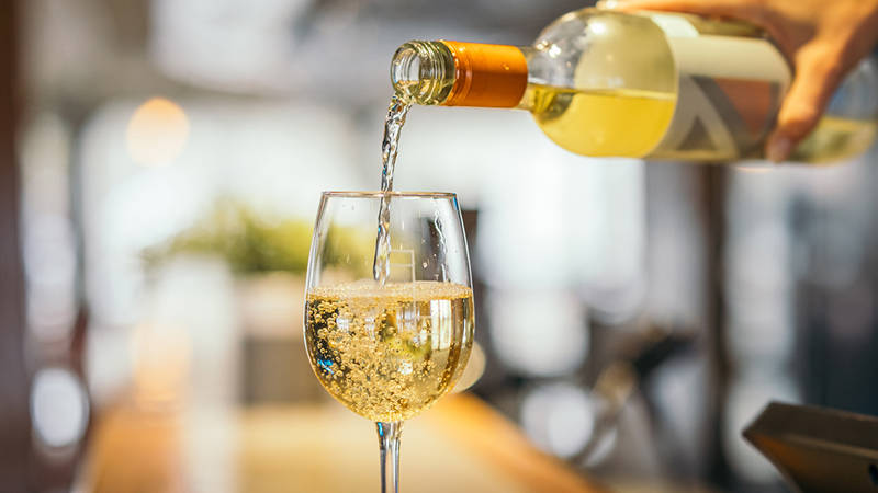 Un gros plan montre une main versant du vin blanc d'une bouteille dans un verre à vin. Le vin pétillant s'écoule doucement et remplit le verre, tandis que l'arrière-plan flou évoque une atmosphère chaleureuse et conviviale