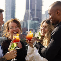 A group of four smiling friends toasting with colorful cocktails on a terrace, with skyscrapers in the background.