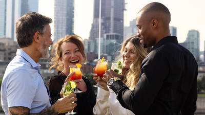 Un groupe de quatre amis souriants trinque avec des cocktails colorés sur une terrasse, avec des gratte-ciel en arrière-plan.