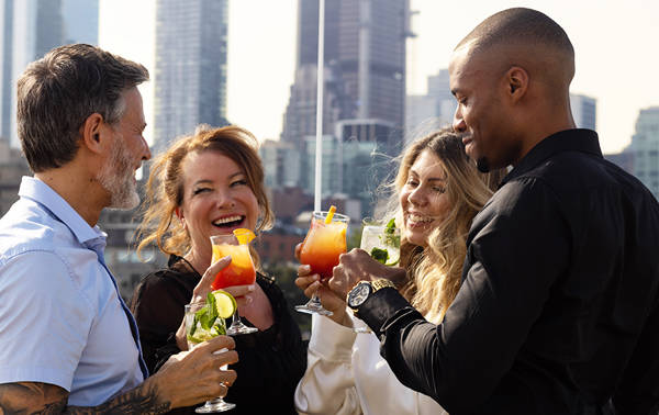 Un groupe de quatre amis souriants trinque avec des cocktails colorés sur une terrasse, avec des gratte-ciel en arrière-plan.