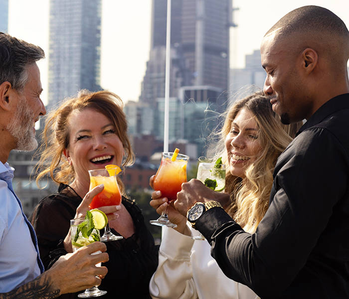 Un groupe de quatre amis souriants trinque avec des cocktails colorés sur une terrasse, avec des gratte-ciel en arrière-plan.