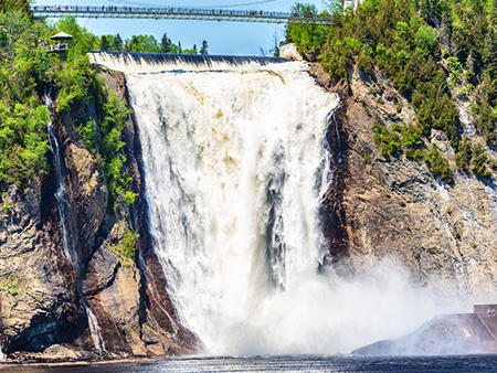 Montmorency Falls in the summer cascading into the Montmorency River below, surrounded by trees with a suspended footbridge above